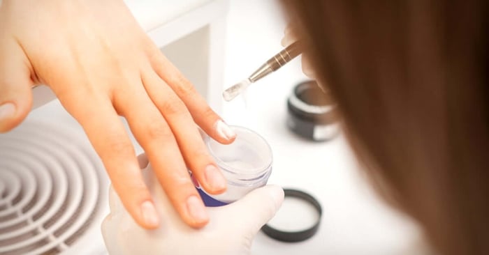 A manicurist holds their client's finger and applies acrylic onto their nail with a small brush.