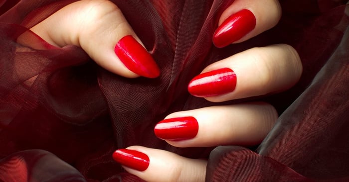 A close-up of a hand pose, showing off solid red nails. The person holds a red fabric to accentuate the pose.