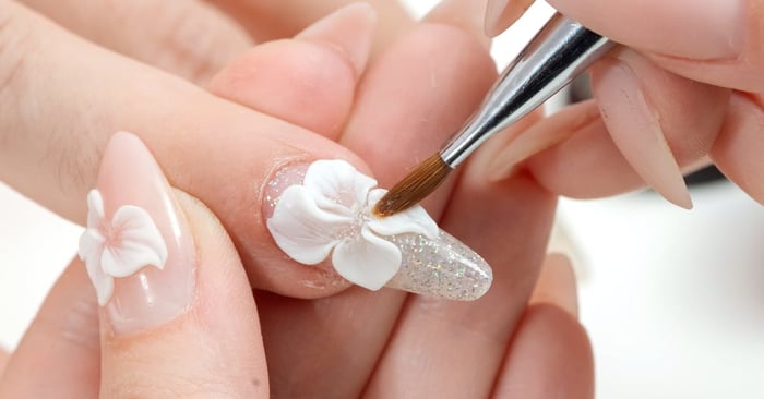 A close-up of someone's pointer finger nail. The nail has silver glitter while someone adds a 3D flower design.