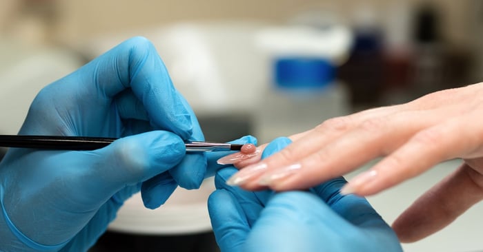 A nail technician in blue gloves applies gel nail polish with a brush to the client's fingernail in a professional salon.
