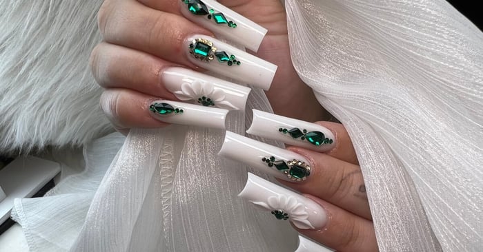 Long white 3D acrylic nails with emerald rhinestones, petal accents, and crystal embellishments on a white tulle backdrop.