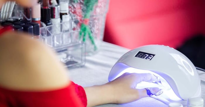A client wearing nail forms and holding one hand beneath a UV curing device with a countdown displayed on top.
