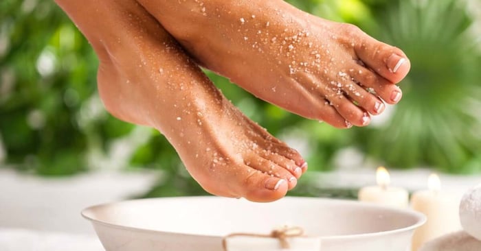 Two feet with bright white French tips sitting above a bowel. The feet have remnants of a sugar scrub across the skin.