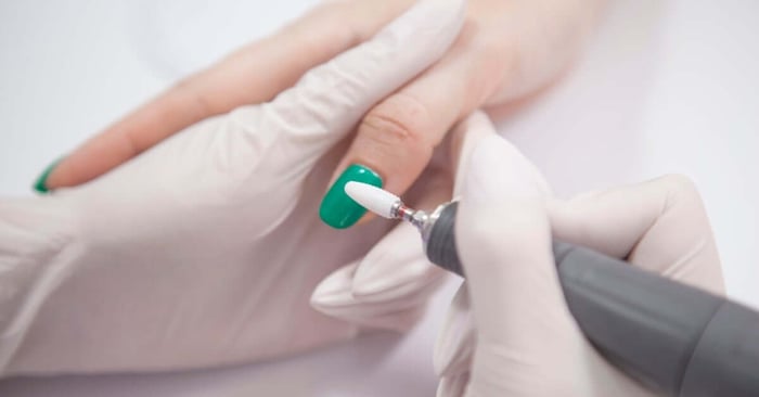 A nail technician in gloves preparing to use a tool to file off the cured green gel coating a client's thumb nail.