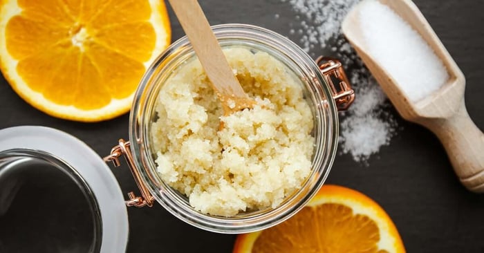 A glass jar containing a light orange sugar scrub. There are orange slices and a scoop of sugar set beside it.