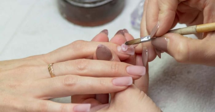 A nail technician with mauve nails holding a client's finger in place while applying a bead of acrylic to their nail.
