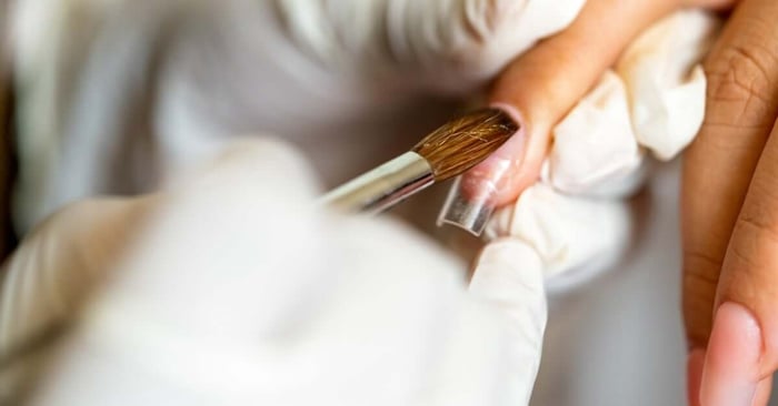 A nail technician holding a client's finger in place and brushing along their nail's surface with a thick, damp brush.
