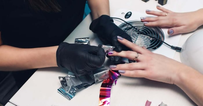 A nail technician wearing black gloves while applying a dual-color foil onto a client's pre-prepped nails.