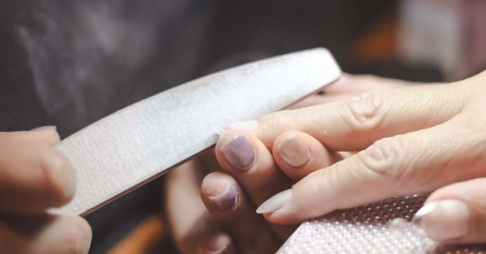 A nail technician using a large file to correct the shape of a client's nails, creating filing dust between them both.