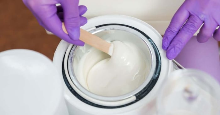A technician in purple gloves stirs white cream wax with a wooden spatula in an open wax warmer.