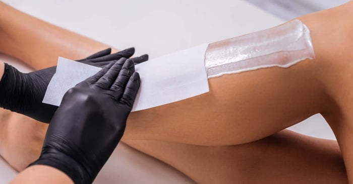 An esthetician in black gloves applies a waxing strip to a client's leg during professional hair removal treatment.