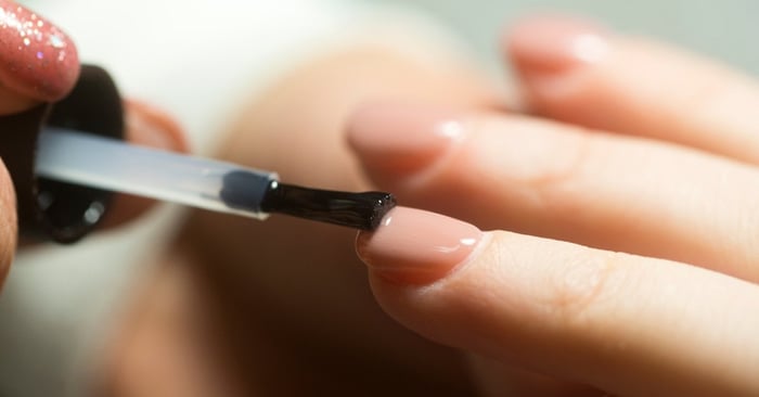 A close-up of a nail tech wearing glitter nail polish painting a client's nail with a clear, natural colored polish.
