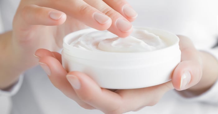 A person in a white long-sleeve shirt applies moisturizing cream from a small white round jar to their hands.
