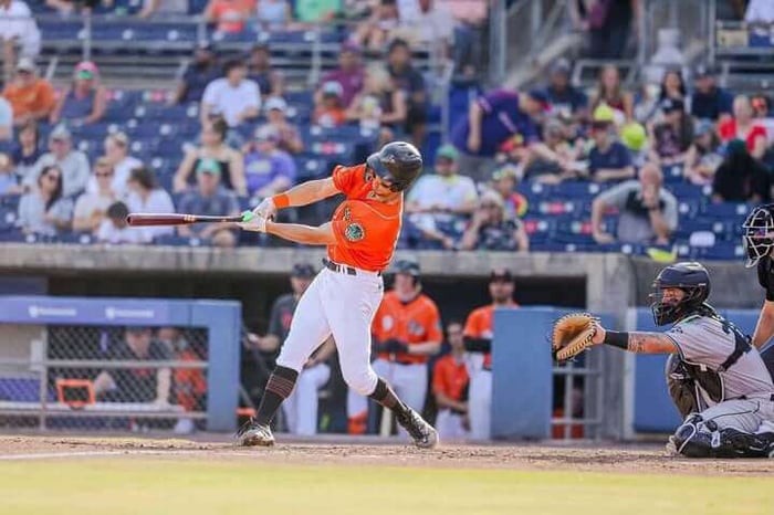 A baseball player in an orange jersey uses a baseball thumb guard as he swings at a pitch, with a catcher and umpire behind and spectators watching from the stands.