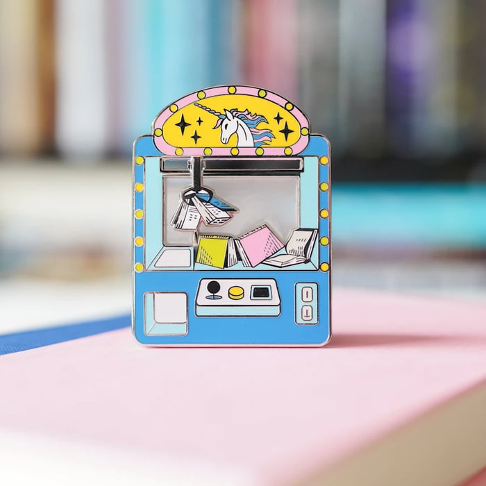his image shows a colorful, whimsical pin depicting a unicorn-themed vending machine. The vending machine is illustrated with a blue and yellow color scheme, featuring a unicorn head at the top with a pink and yellow halo. Inside the vending machine, there are colorful books and a small figure holding a book, suggesting the vending machine dispenses books. The machine has a joystick and buttons, adding to the playful design. The pin is placed on a pink surface, possibly a notebook or a piece of paper, with a blurred background of what appears to be a bookshelf filled with books.