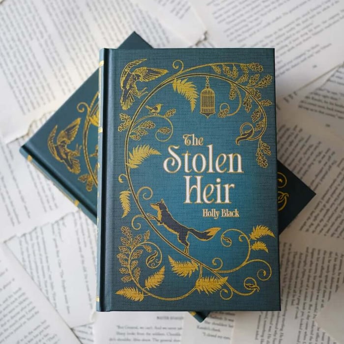 This image shows a close-up view of two hardcover books stacked on top of each other, placed on a surface covered with open pages of other books. The top book has a teal cover with an intricate golden design featuring leaves, birds, and a fox. The title "The Stolen Heir" is prominently displayed in large, golden letters, with the author's name "Holly Black" beneath it. The bottom book also has a teal cover with similar golden designs and the same title and author's name. The background consists of various pages of text, some of which are visible and partially legible.