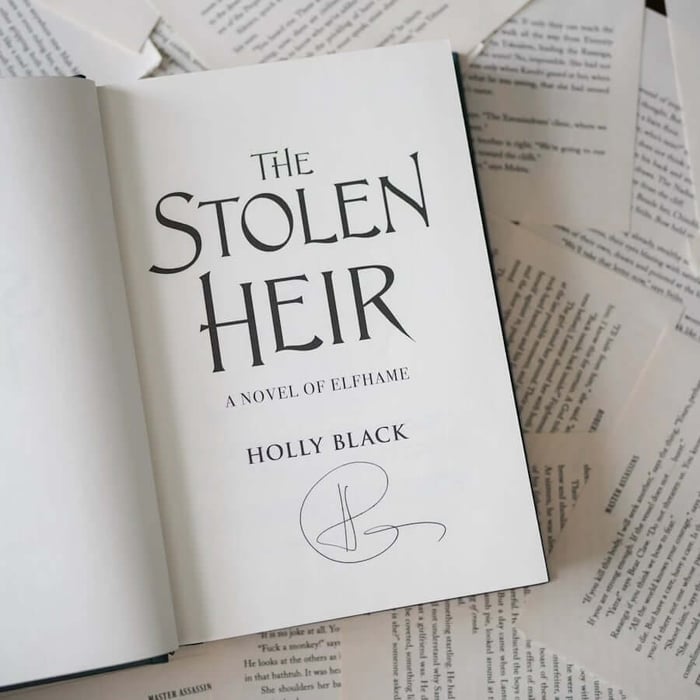 This image shows a book titled "The Stolen Heir" by Holly Black, which is open to a page with handwritten text. The book appears to be placed on top of several other books, with their pages visible in the background. The handwritten text on the page is partially visible and seems to be a continuation of the story. The book cover features a simple design with the title and author's name prominently displayed.