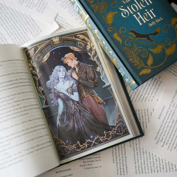 This image shows an open book displaying a detailed illustration of a man and a woman in a romantic embrace. The book is placed on a surface with other papers and books visible in the background. The illustration is framed with ornate, golden designs. The book's cover, partially visible in the top right corner, features the title "The Stolen Heir" by Holly Black, with a fox logo and golden floral patterns. The pages of the book contain text, with visible words such as "golden," "invited," and "halls," suggesting a narrative context.