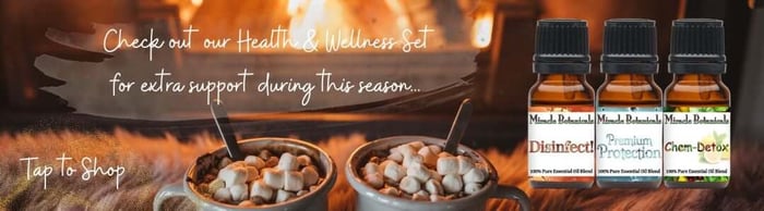 Two cups of hot chocolate in front of a warm fireplace, with three amber bottles of essential oil blends by Miracle Botanicals, including Disinfect blend, Premium Protection blend, and Chem-Detox blend. Text reads "Check out our Health and Wellness Set for extra support during this season... Tap to Shop"