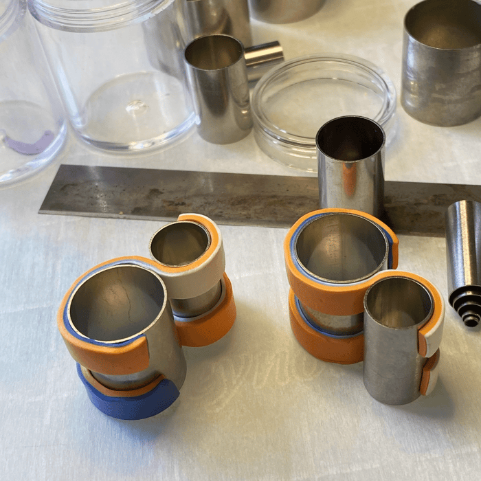 showing a multicolored strip of clay wound in one direction around one circular metal form and in the opposite direction around another.