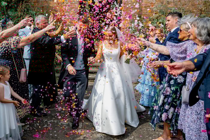 A groom and bride walk through a wedding confetti storm of pinks and oranges. Their guests are smiling as they throw the wedding confetti.