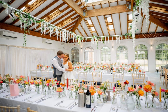 A groom kisses his bride in an airy room, decorated for the wedding breakfast with pink and orange flowers.