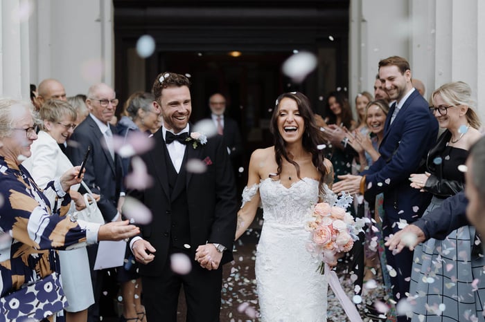 A bride and groom laugh as they walk through a wedding confetti cloud.