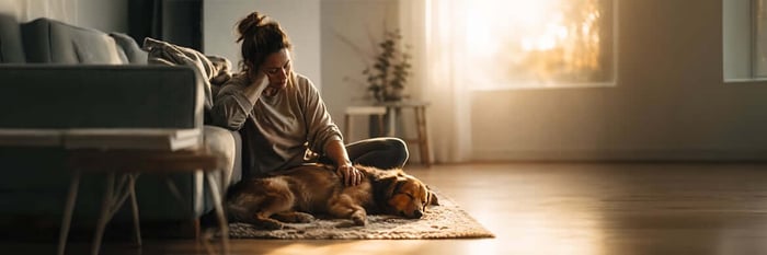 A woman sits on the floor leaning against a couch, head resting on one hand, her other hand on a dog asleep in her lap as low evening light comes through the window.