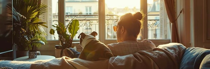 Young professional unwinding on the sofa with their dog, sharing a quiet golden-hour moment in a cozy, plant-filled city apartment.