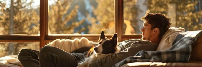 Young man resting on a cozy cabin sofa beside his black and white dog, both calm and content in the golden hour light—feel better, THiNK LiKE A DOG.
