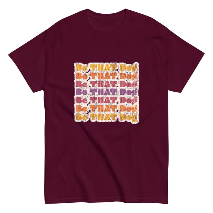 Unisex maroon t-shirt featuring a retro-inspired design with the repeated phrase 'Be THAT Dog' in a distressed font. The text alternates in vibrant shades of orange, purple, and white, creating a bold and playful look for dog lovers.