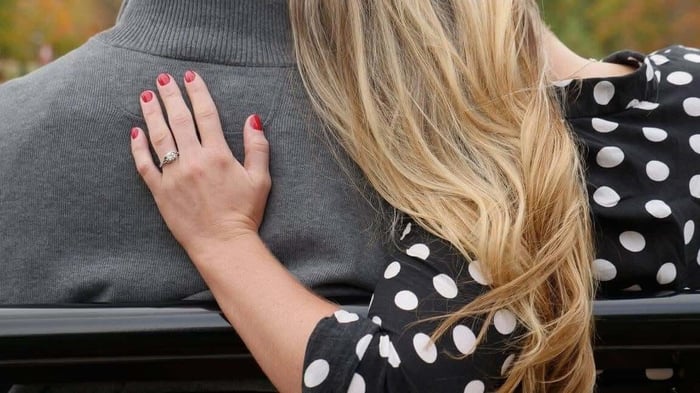 Two people sitting on a park bench with arms around each other, looking out at autumn scenery.