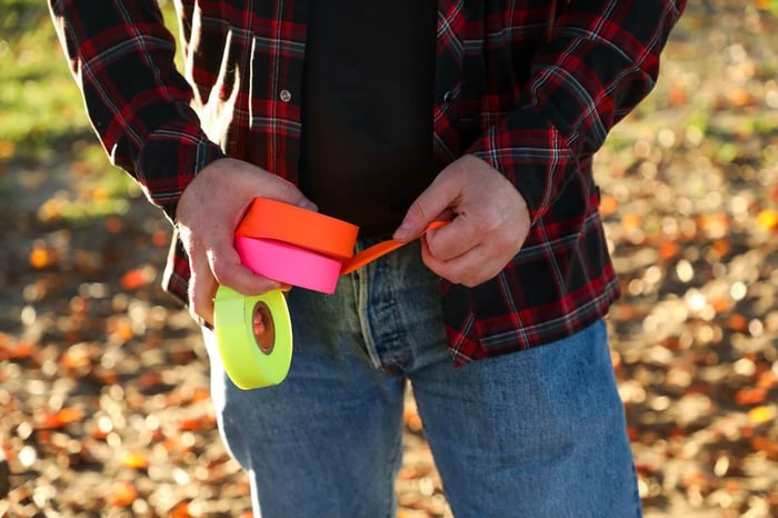 man unraveling orange flagging tape while holding pink and yellow flagging tapes