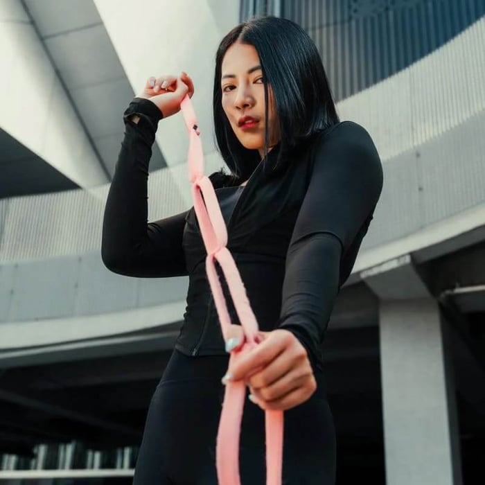 一名穿著黑色運動服的女性雙手拉伸粉紅色多功能阻力彈力帶，在室內進行局部線條雕塑的基礎居家運動。