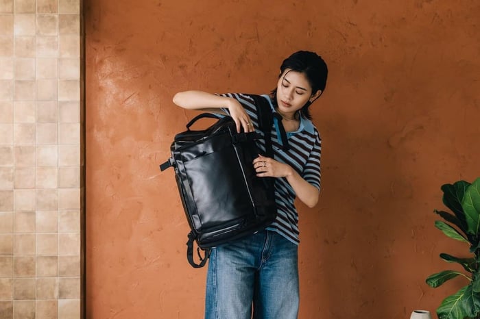 女性模特兒率性手提黑色 Bagsmart 旅人機能擴充後背包 38L(Pro-Tech 強化款),展現高階 TPU 覆膜面料的霧面高質感與俐落外觀,完美呼應 PTT 與 Dcard 網友對於商務出差與隨身登機「旅行後背包」的時尚穿搭與「女後背包推薦」美學需求。