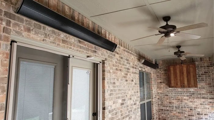 Two sleek, black wall-mounted electric patio heaters installed high on a brick patio wall, with ceiling fans visible overhead.