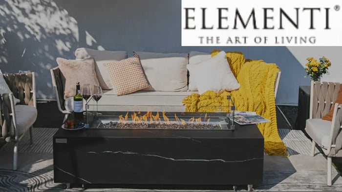 Elementi fire pit table with flames, set on a patio with a couch and chairs.