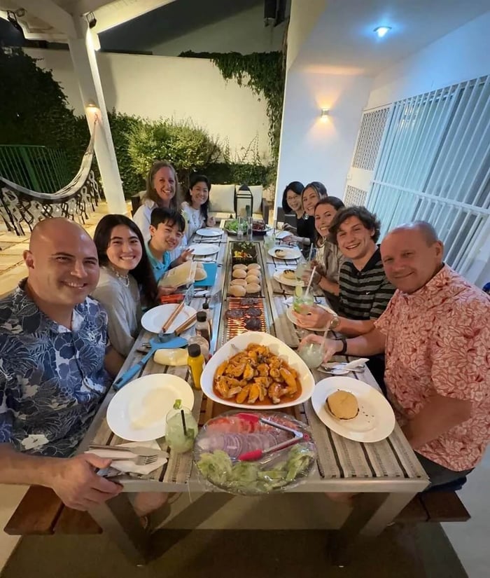  A diverse group of family and friends is gathered around an Angara BBQ table for an outdoor dinner. The table is laden with various dishes, including a large bowl of grilled shrimp in the center. 