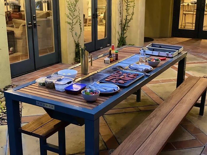 An Angara BBQ table is set up on a patio, ready for an outdoor meal. The long, modern table features a slatted wood top and a built-in grill in the center, which holds a pan of raw meat and other items ready for cooking.