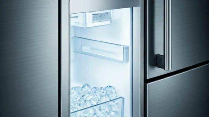A close-up of a stainless steel refrigerator door with an open ice dispenser. Inside, a bin is filled with a large quantity of crescent-shaped ice cubes, glistening under a bright light. The dispenser appears to be a built-in feature of the refrigerator.