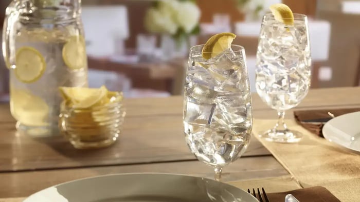 A close-up of two tall glasses filled with ice cubes and water on a dining table. The glasses have a lemon slice garnish. A pitcher filled with water and lemon slices is visible in the background. The table is set with plates and cutlery.