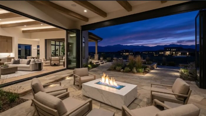 A wide-angle, luxury architectural photograph taken at dusk from inside an open-concept living room, looking out onto a large, raw stone patio. Expansive 15-foot bi-fold glass doors are fully retracted to create a seamless indoor-outdoor transition. On the flagstone terrace, a modern, rectangular Elementi Carrara fire pit table made of white marble porcelain sits center stage, featuring a vibrant linear flame surrounded by blue fire glass. High-end outdoor armchairs surround the fire feature, while the background reveals a beautifully landscaped yard with native grasses and a stunning twilight view of distant rolling mountains under a deep blue and purple sunset sky.