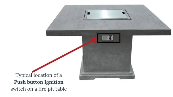 A concrete square fire pit table with a closed burner cover. A red arrow points to a rectangular push-button ignition switch located on the side of the table's base.