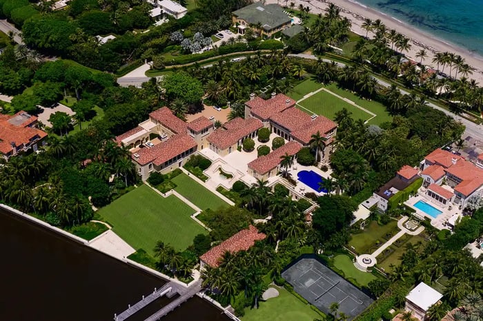 An aerial view of a large, Mediterranean-style mansion on the waterfront. The property features a tennis court, swimming pool, and lush green lawns. The mansion is surrounded by other houses and palm trees.