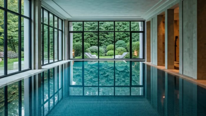 A tranquil indoor swimming pool, bathed in natural light from floor-to-ceiling windows, offering a serene view of a landscaped garden.