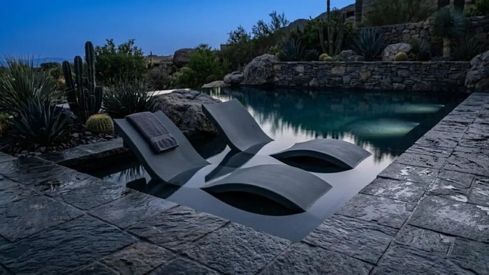 A dramatic blue hour photograph of a luxury dark bottom pool in a US backyard. Two Granite Grey Autograph in pool loungers are submerged in a deep navy pool ledge surrounded by charcoal flagstone and basalt rock. The image illustrates the mirror like reflection of the sky and the stone like matte finish of the furniture for a biophilic retreat by Lux Yard and Homes.