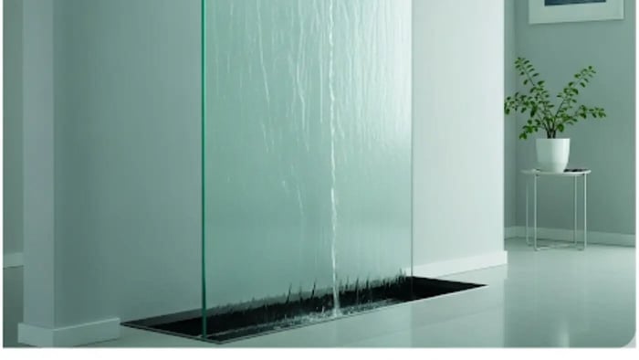 A stunning indoor wall water feature made of clear glass with a sleek, modern design. Water cascades down the smooth surface, with lights reflecting off the glass and the flowing water.
