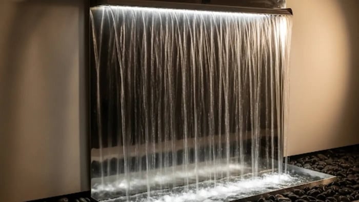 A serene indoor wall fountain with water flowing in a thin, continuous curtain over a polished metal sheet, illuminated from above to create a mesmerizing visual effect.