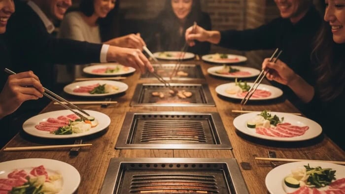 A group of friends gathered around a long, rectangular wooden Korean BBQ table. The table features multiple recessed grill units down the center, allowing guests on both sides to use tongs to flip meat and vegetables directly from their seats. Each person has a white plate with various raw meats and side dishes, illustrating the interactive, communal dining experience of social grilling.