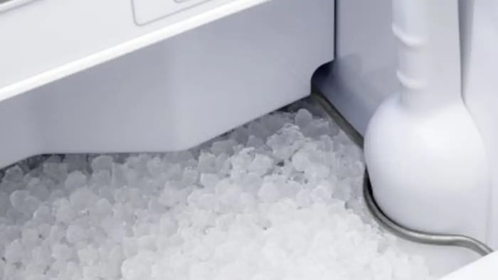 A close-up view of an ice maker bin. The bin is filled with a large quantity of crescent-shaped ice cubes. A white plastic scoop is visible in the bin, used to dispense the ice. The ice cubes are glistening and appear to be freshly made.